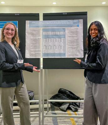 普拉提亚·普萨拉和萨拉·曼站在一张海报前，海报上记录了一项名为“基于特质的正念和DBT情绪调节技能的使用:新冠肺炎初期封锁期间的一年级大学生”的研究项目.